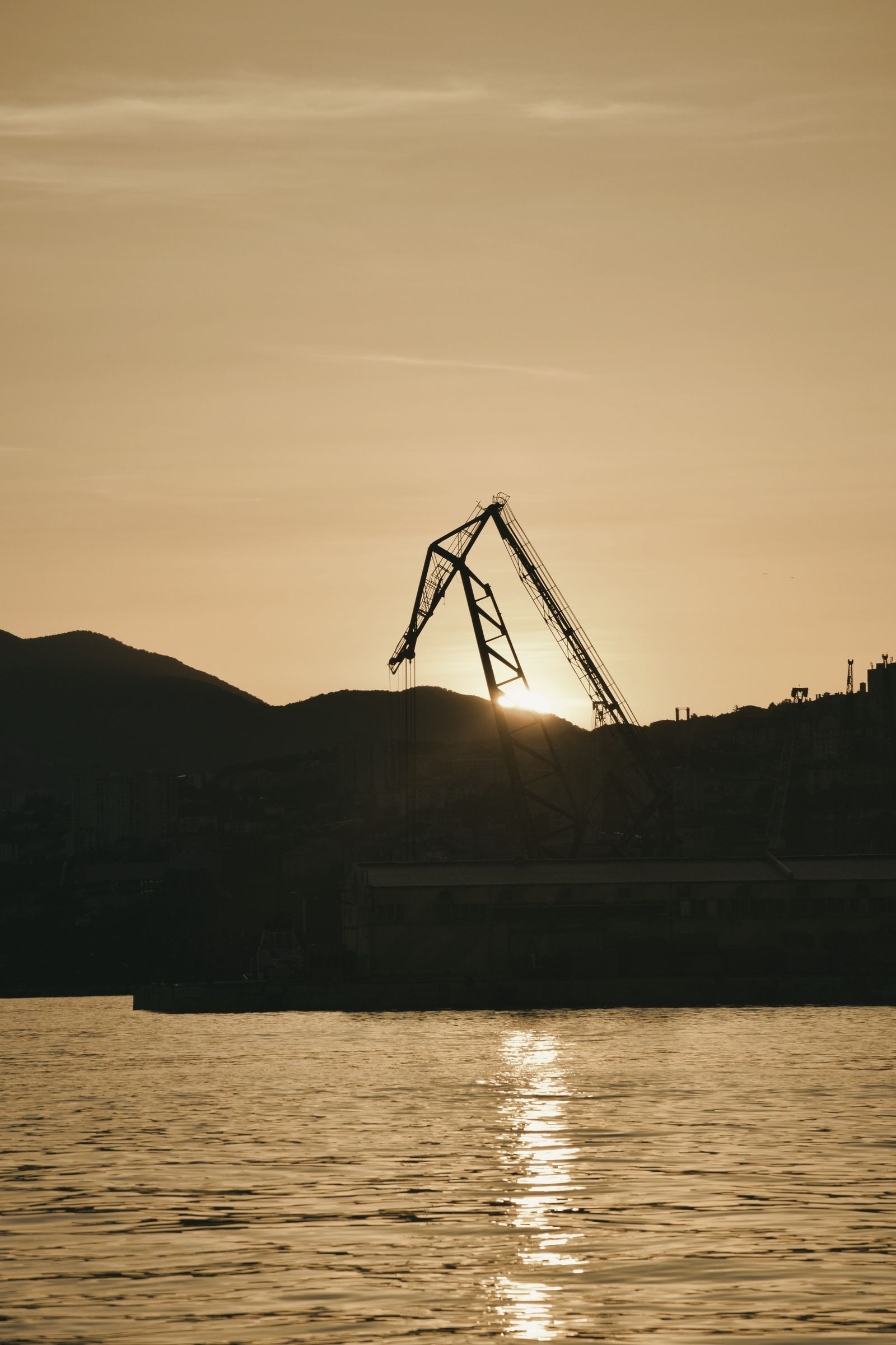 O macara portuară în contralumină, cu soarele jos la apus și apa liniștită dedesubt